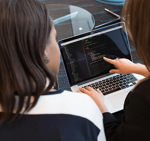 "Two women collaborating on laptop, writing code in a tech workspace"